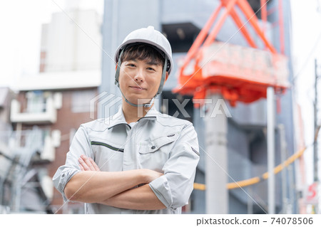 一名戴著頭盔和雙臂交叉在建築工地的年輕工人 74078506