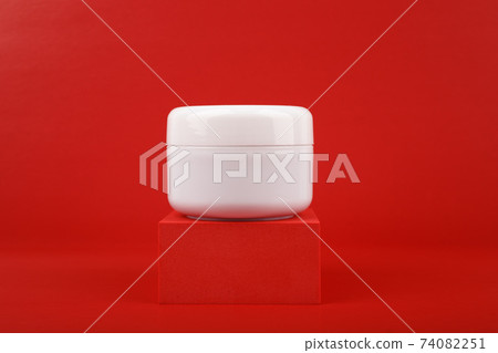 Still life with close up of face cream. mask or balm in white jar on red square shaped podium against red background 74082251