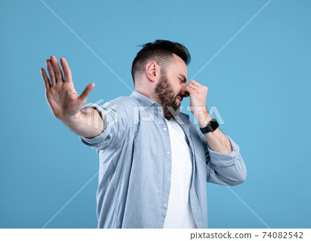 Bad smell. Young bearded guy closing his nose and showing STOP gesture, expressing disgust on blue studio background Bad smell. Young bearded guy closing his nose and showing STOP gesture, expressing disgust on blue studio background 74082542