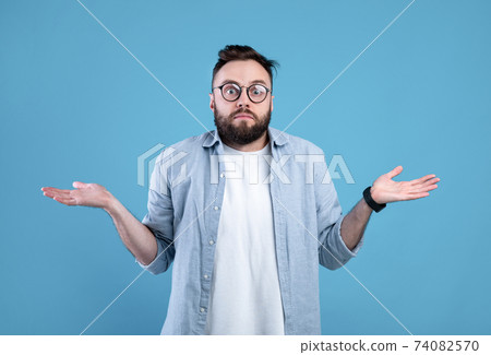 Who knows. Confused bearded man in glasses shrugging shoulders on blue studio background Who knows. Confused bearded man in glasses shrugging shoulders on blue studio background 74082570