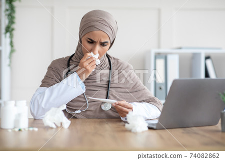 Sick muslim woman doctor sneezing nose and looking at thermometer Sick muslim woman doctor sneezing nose and looking at thermometer 74082862
