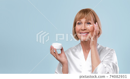 Smiling mature woman applying rejuvenating cream on her face over blue studio background, banner design with empty space 74082931