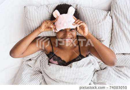 Happy young african american attractive female taking off pink sleep mask have charming smile Happy young african american attractive female taking off pink sleep mask have charming smile 74083050