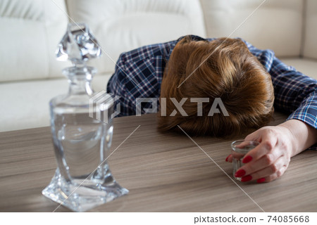 A drunk woman with shot of vodka and a decanter lies face down on the table 74085668