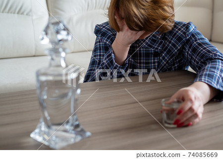 Drunk woman with a glass of vodka and a decanter covers her face with her hand Drunk woman with a glass of vodka and a decanter covers her face with her hand 74085669