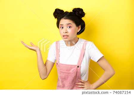 Confused asian teen girl with bright makeup, raising hand up clueless, looking puzzled, cant understand something, pointing at copy space, yellow background Confused asian teen girl with bright makeup, raising hand up clueless, looking puzzled, cant understand something, pointing at copy space, yellow background 74087154