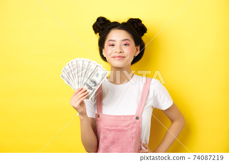 Shopping. Smiling asian girl with stylish makeup, showing dollar bills with pleased face, making money, standing on yellow background Shopping. Smiling asian girl with stylish makeup, showing dollar bills with pleased face, making money, standing on yellow background 74087219