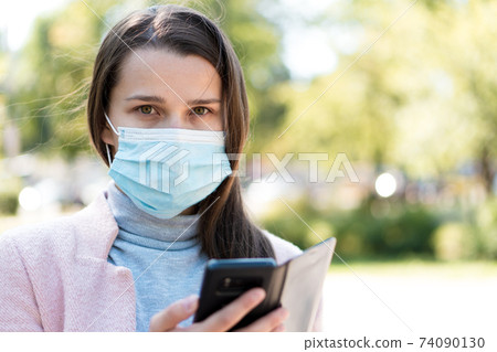 care, infants, spring, cvid-19, quarantine concept - Young cute long haired woman European Caucasian Slavic appearance in medical protective mask uses smartphone in midday sunlight backlight in park 74090130