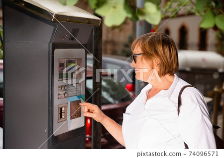 Smiling middle aged woman using parking machine to pay for car parking on summer city street Smiling middle aged woman using parking machine to pay for car parking on summer city street 74096571