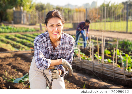 Woman gardener holding shovel in garden against background of working people Woman gardener holding shovel in garden against background of working people 74096623