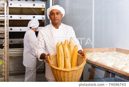 Male baker holding basket of hot baguettes in the kitchen of bakery Male baker holding basket of hot baguettes in the kitchen of bakery 74096743