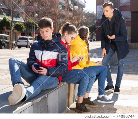 Four teenagers enthusiastically look at the screens of their smartphones on street Four teenagers enthusiastically look at the screens of their smartphones on street 74096744