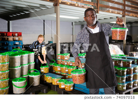 Afro-american male worker stocks plastic containers and cans with olives in warehouse Afro-american male worker stocks plastic containers and cans with olives in warehouse 74097018