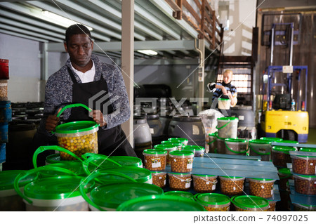 Afro-american male worker stocks plastic containers and cans with olives in warehouse Afro-american male worker stocks plastic containers and cans with olives in warehouse 74097025