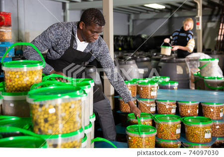 Afro-american male worker stocks plastic containers and cans with olives in warehouse Afro-american male worker stocks plastic containers and cans with olives in warehouse 74097030