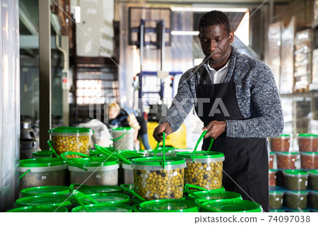 Afro-american male worker stocks plastic containers and cans with olives in warehouse Afro-american male worker stocks plastic containers and cans with olives in warehouse 74097038