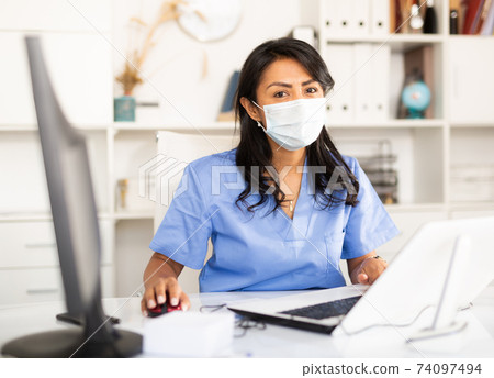 Woman general practitioner in mask working on laptop in office 74097494