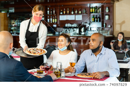 Waitress in protective mask serving delicious pizza to friends in restaurant Waitress in protective mask serving delicious pizza to friends in restaurant 74097829