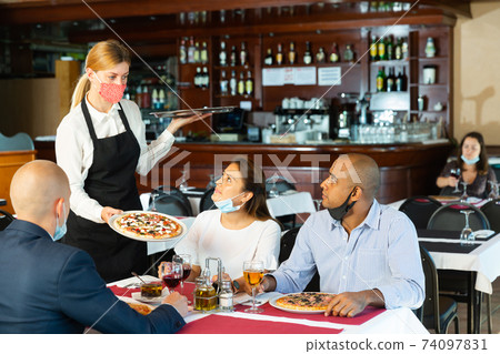 Polite waitress in protective mask bringing ordered pizza to friends visited restaurant Polite waitress in protective mask bringing ordered pizza to friends visited restaurant 74097831
