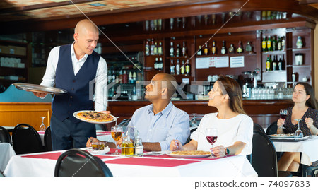 Polite waiter serving pizza to hispanic couple in restaurant Polite waiter serving pizza to hispanic couple in restaurant 74097833