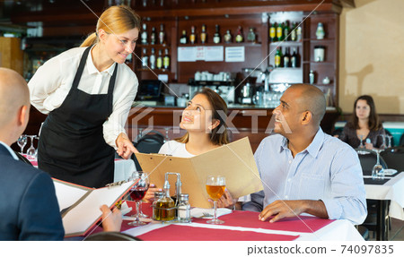 Restaurant guests will choose dish from the menu. Polite waitress helps with choice 74097835