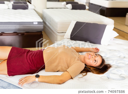 Smiling Hispanic girl lying on new modern orthopedic mattress in furniture shop trying its comfort and convenience Smiling Hispanic girl lying on new modern orthopedic mattress in furniture shop trying its comfort and convenience 74097886