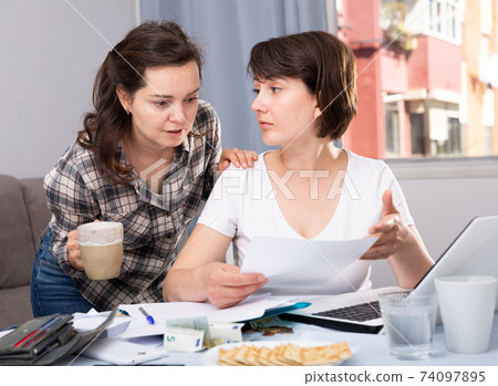 Two smiling women sitting at home table with laptop Two smiling women sitting at home table with laptop 74097895