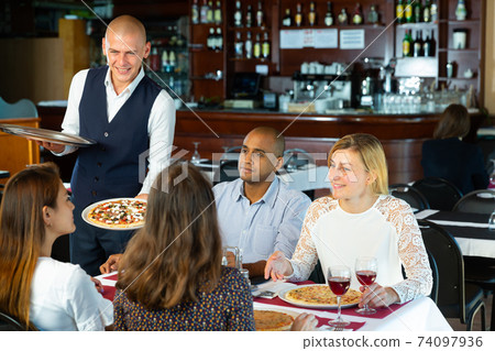 Polite waiter serving pizza to friendly company in pizzeria Polite waiter serving pizza to friendly company in pizzeria 74097936