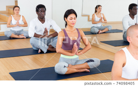 Asian woman meditating in yoga position Padmasana with group at gym Asian woman meditating in yoga position Padmasana with group at gym 74098405