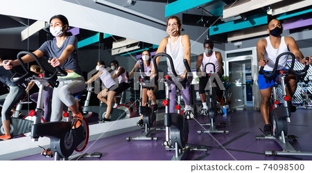 Men and women wearing protective masks ride stationary bike in fitness club Men and women wearing protective masks ride stationary bike in fitness club 74098500