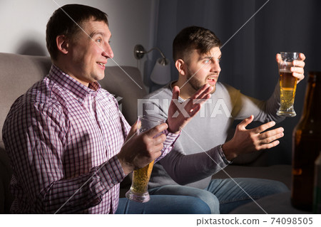 Two happy men emotionally watching tv at home Two happy men emotionally watching tv at home 74098505