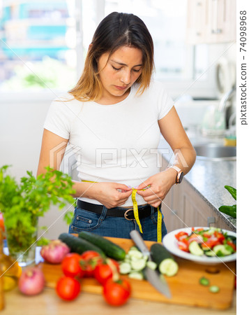 Upset housewife measures waist before eating vegetable salad Upset housewife measures waist before eating vegetable salad 74098968