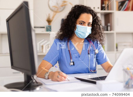 Female therapist in protective mask working at table with laptop in office Female therapist in protective mask working at table with laptop in office 74099404