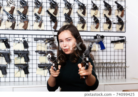 Positive young woman with two revolvers in hands in gun shop Positive young woman with two revolvers in hands in gun shop 74099425