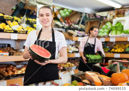 Young woman seller holding half of watermelon in hands in fruit store Young woman seller holding half of watermelon in hands in fruit store 74099535