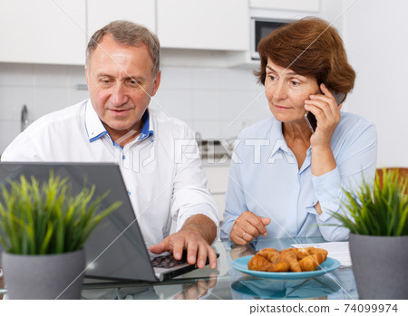 Smiling mature family couple with phone using laptop at kitchen table Smiling mature family couple with phone using laptop at kitchen table 74099974
