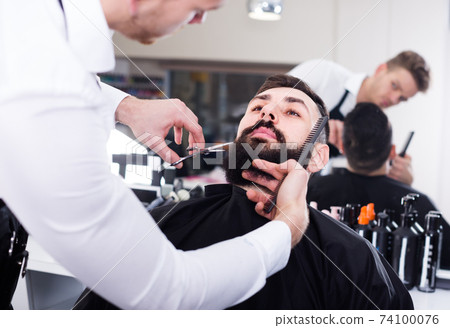 Young man forming beard of client into shape Young man forming beard of client into shape 74100076