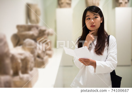 Chinese woman visitor with guide book looking at exhibition in museum Chinese woman visitor with guide book looking at exhibition in museum 74100687