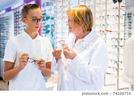 Mature female ophthalmologist helping teen boy to choose glasses Mature female ophthalmologist helping teen boy to choose glasses 74100759