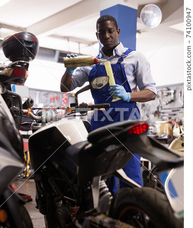 Engineer in overalls pours oil for motorcycle engine in garage Engineer in overalls pours oil for motorcycle engine in garage 74100947