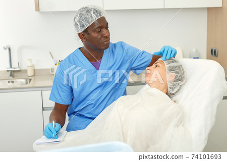 Male examining patient face skin before aesthetic procedure, writing to workbook Male examining patient face skin before aesthetic procedure, writing to workbook 74101693