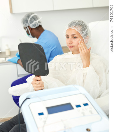 Young woman sitting in armchair and looking to mirror before mesotherapy Young woman sitting in armchair and looking to mirror before mesotherapy 74101702