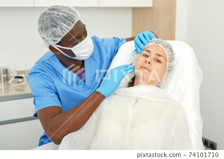 Doctor in mask is doing injection to woman patient in skin of face in estetic clinic 74101710