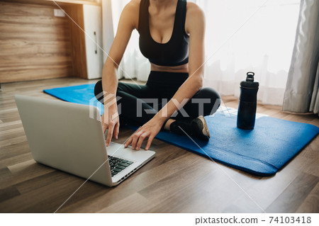 sporty woman in sportswear is sitting on the floor with dumbbells and a protein shake or a bottle of water and is using a laptop. sporty woman in sportswear is sitting on the floor with dumbbells and a protein shake or a bottle of water and is using a laptop. 74103418