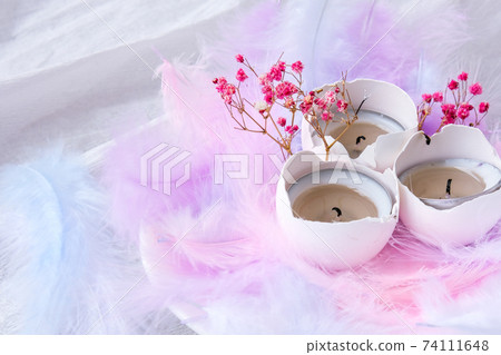 Composition of White egg shell with candle inside. Bright easter decorations with bird feathers. Pastel colors. Easter greeting card. Composition of White egg shell with candle inside. Bright easter decorations with bird feathers. Pastel colors. Easter greeting card. 74111648