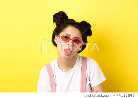 Summer and beauty concept. Childish asian girl in sunglasses showing tongue, being rude, standing on yellow background 74112848