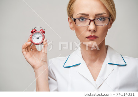 smiley nurse or doctor pointing holding small red alarm clock satanding over grey background 74114331