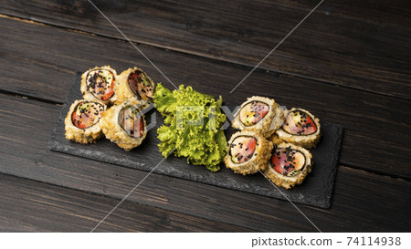 Custom sushi roll in tempura with nori, fresh salmon, tuna, avocado, masago caviar, drizzled with pineapple sauce with salad pouring as decoration on a black plate on a wooden table and background. 74114938