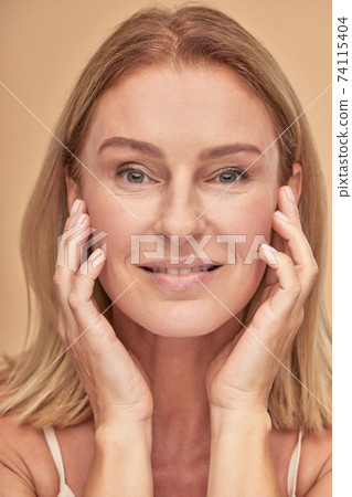 Cosmetology procedures. Attractive middle aged caucasian woman touching her clean and soft skin, looking at camera while posing over beige background in studio Cosmetology procedures. Attractive middle aged caucasian woman touching her clean and soft skin, looking at camera while posing over beige background in studio 74115404