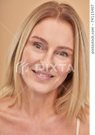 Healthy skin. Close up portrait of a charming happy mature caucasian woman smiling at camera while posing over beige background in studio Healthy skin. Close up portrait of a charming happy mature caucasian woman smiling at camera while posing over beige background in studio 74115407